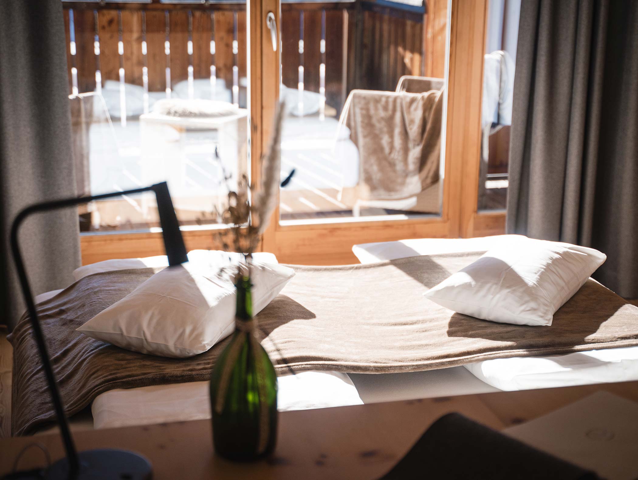 Gemütliches, sonnendurchflutetes Schlafzimmer mit zwei Kissen und einer braunen Decke, in der Nähe eines Fensters mit Blick auf einen Balkon. - Hotel Moosmair