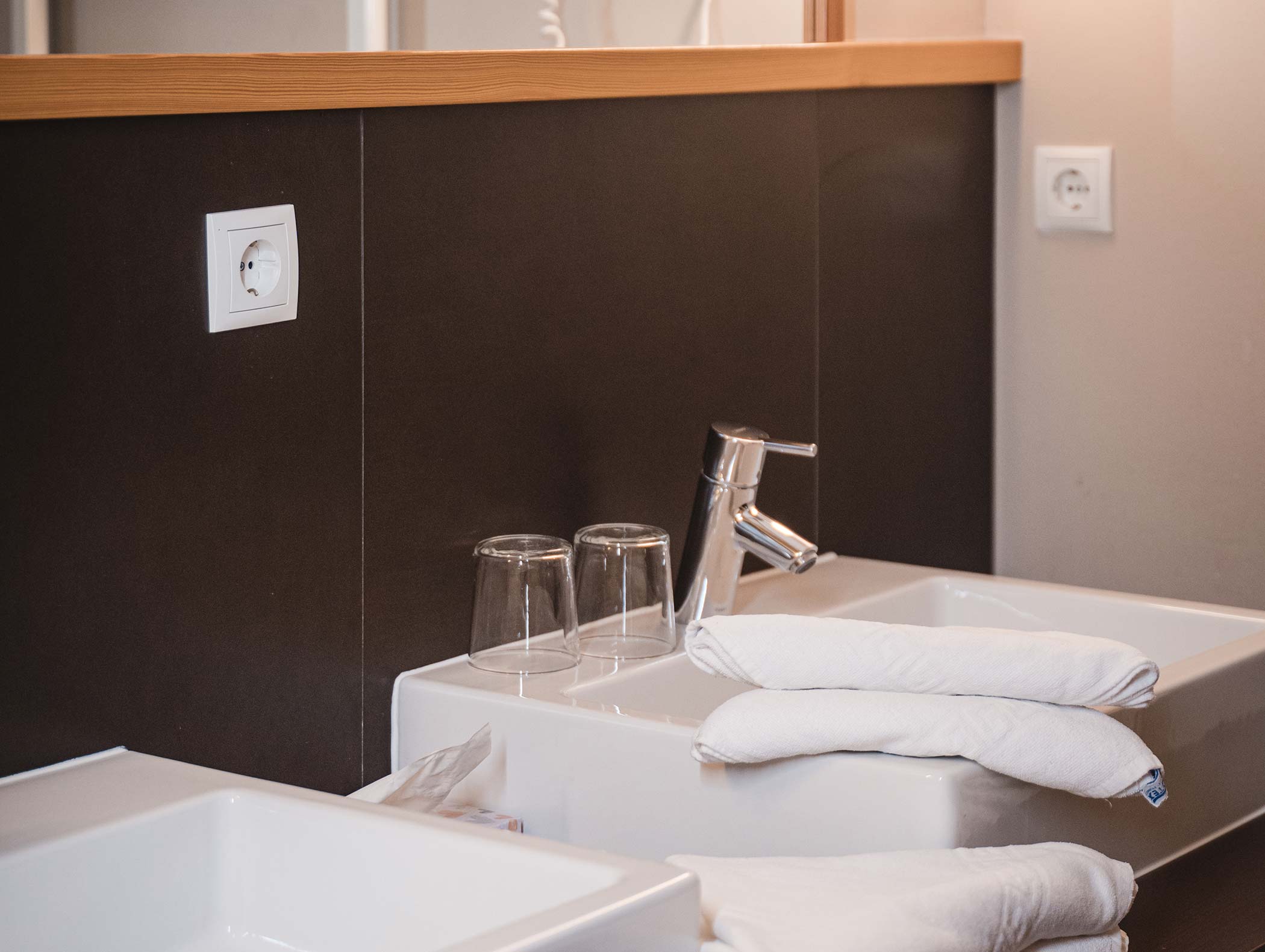 Zwei Waschbecken mit gefalteten weißen Handtüchern und zwei umgedrehten Gläsern auf einem Badezimmertisch neben einer Steckdose. - Hotel Moosmair
