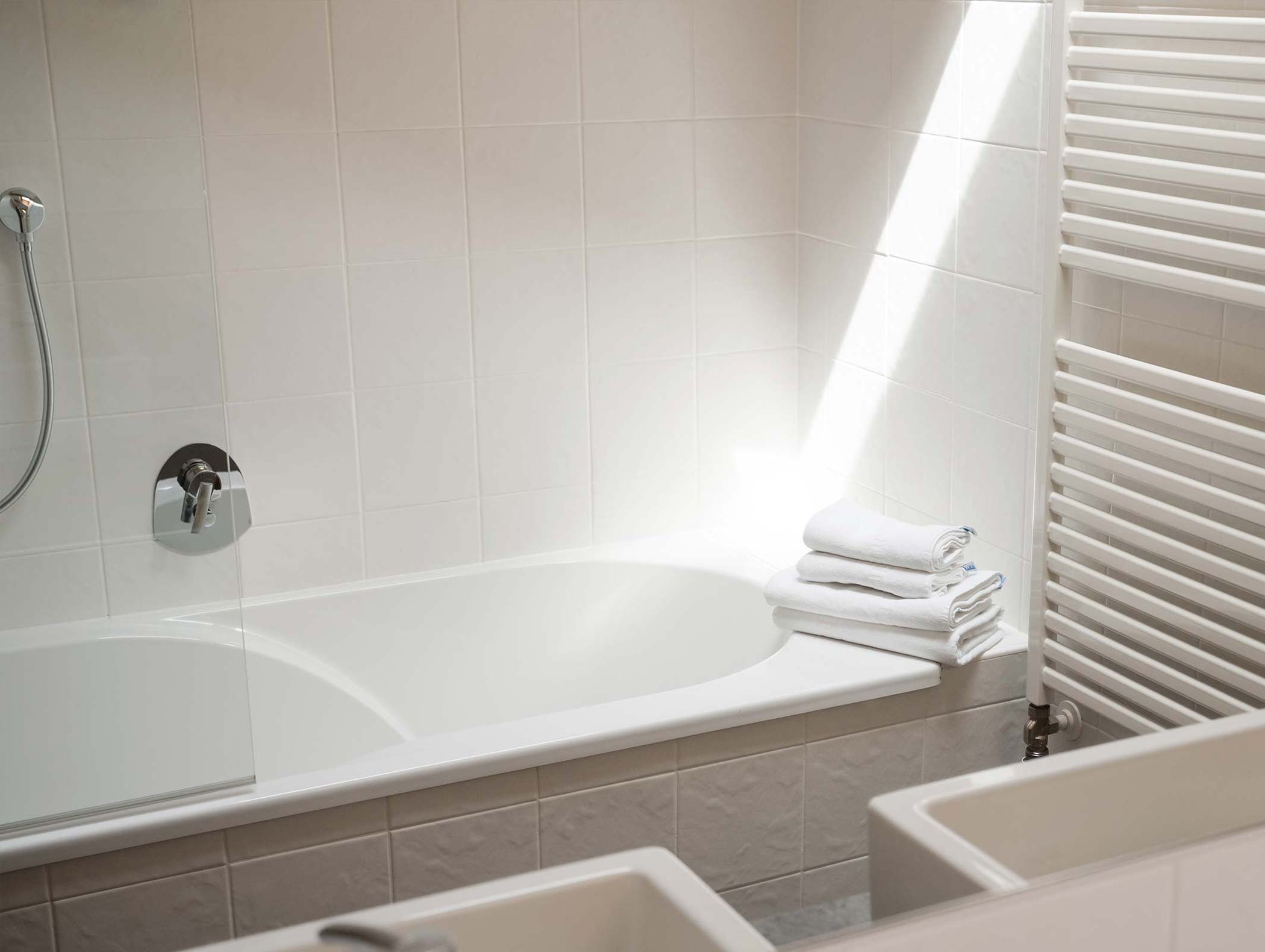 Eine weiße Badewanne mit gefalteten Handtüchern daneben, neben einem Wandheizkörper in einem hellen Badezimmer. - Hotel Moosmair