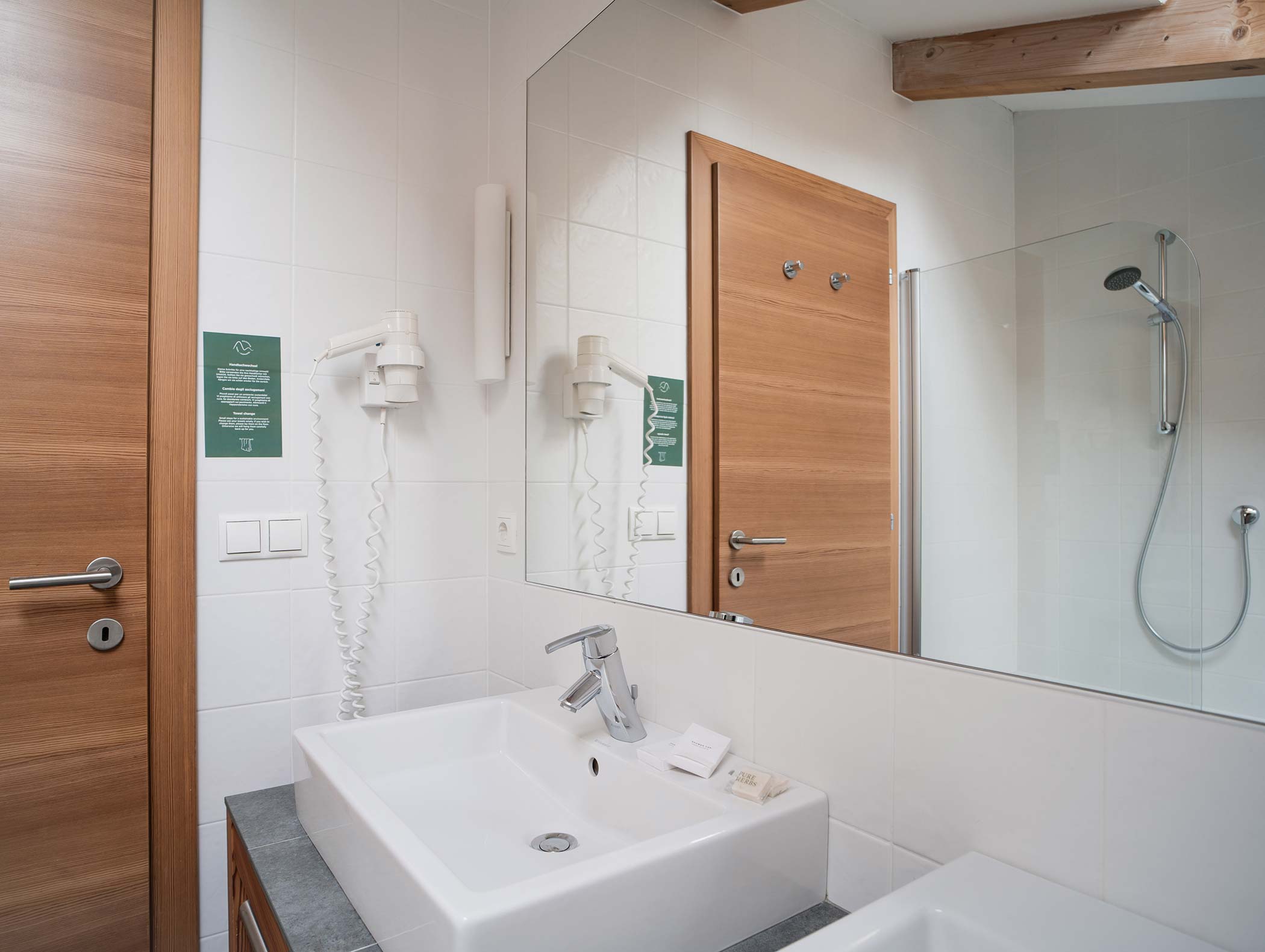 Modernes Badezimmer mit weißem Waschbecken, Wandhaartrockner, Holztüren und Glasduschkabine, die sich in einem Spiegel spiegelt. - Hotel Moosmair