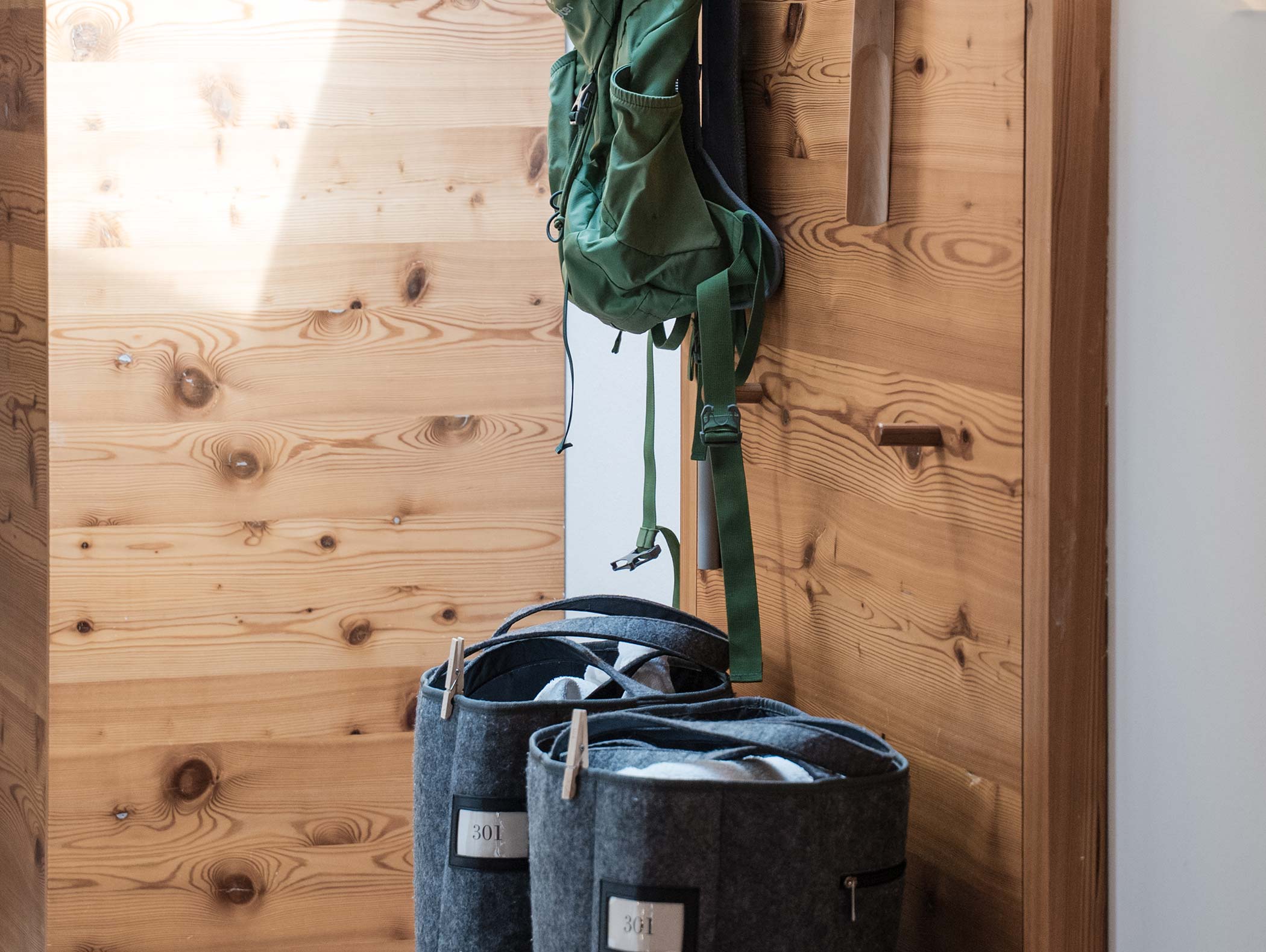 Ein grüner Rucksack hängt an einer Holzwand über zwei grauen Filzkörben mit zusammengerollten Decken darin. - Hotel Moosmair