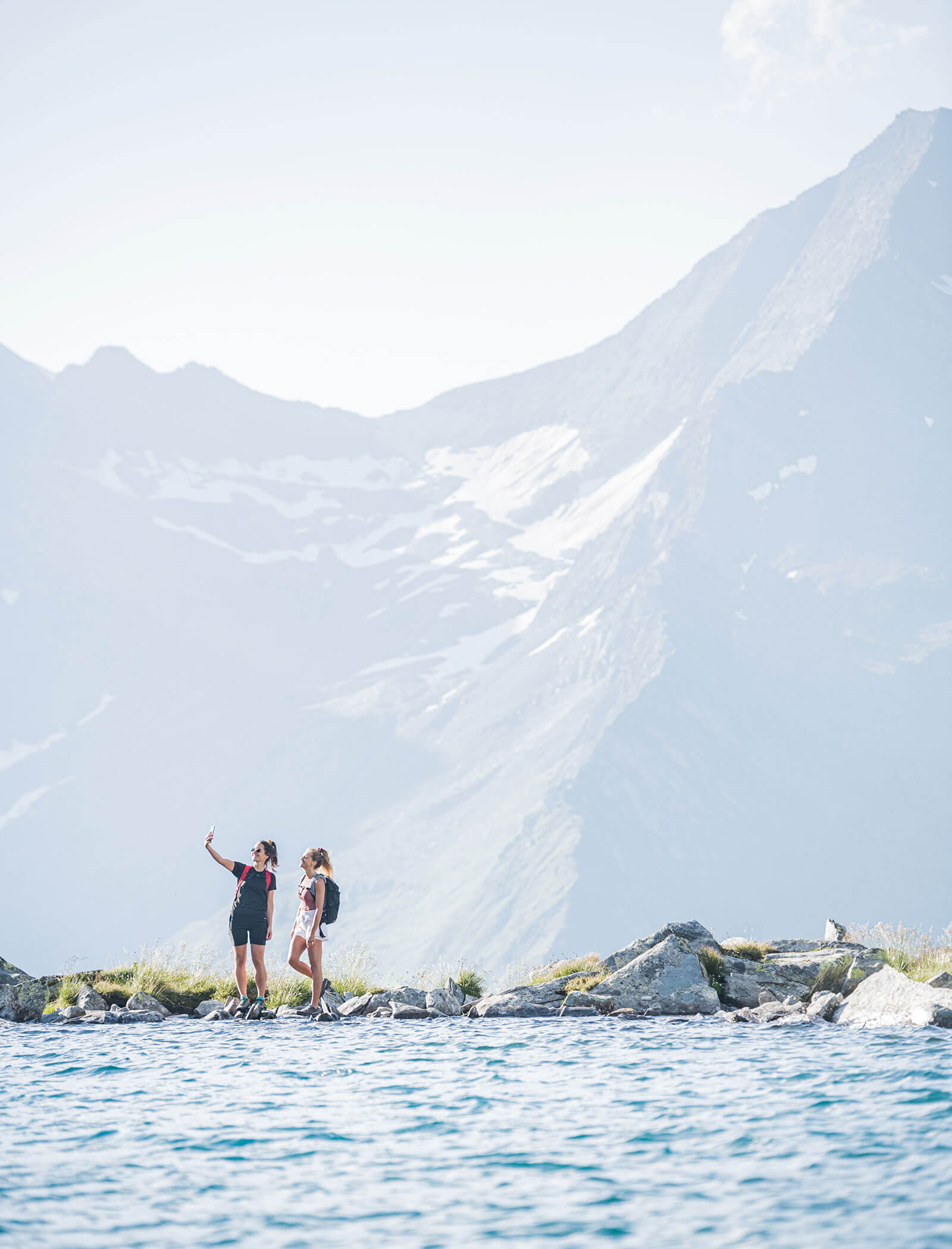 Zwei Wanderer machen ein Selfie an einem Bergsee mit hohen Schneegipfeln im Hintergrund. - Hotel Moosmair