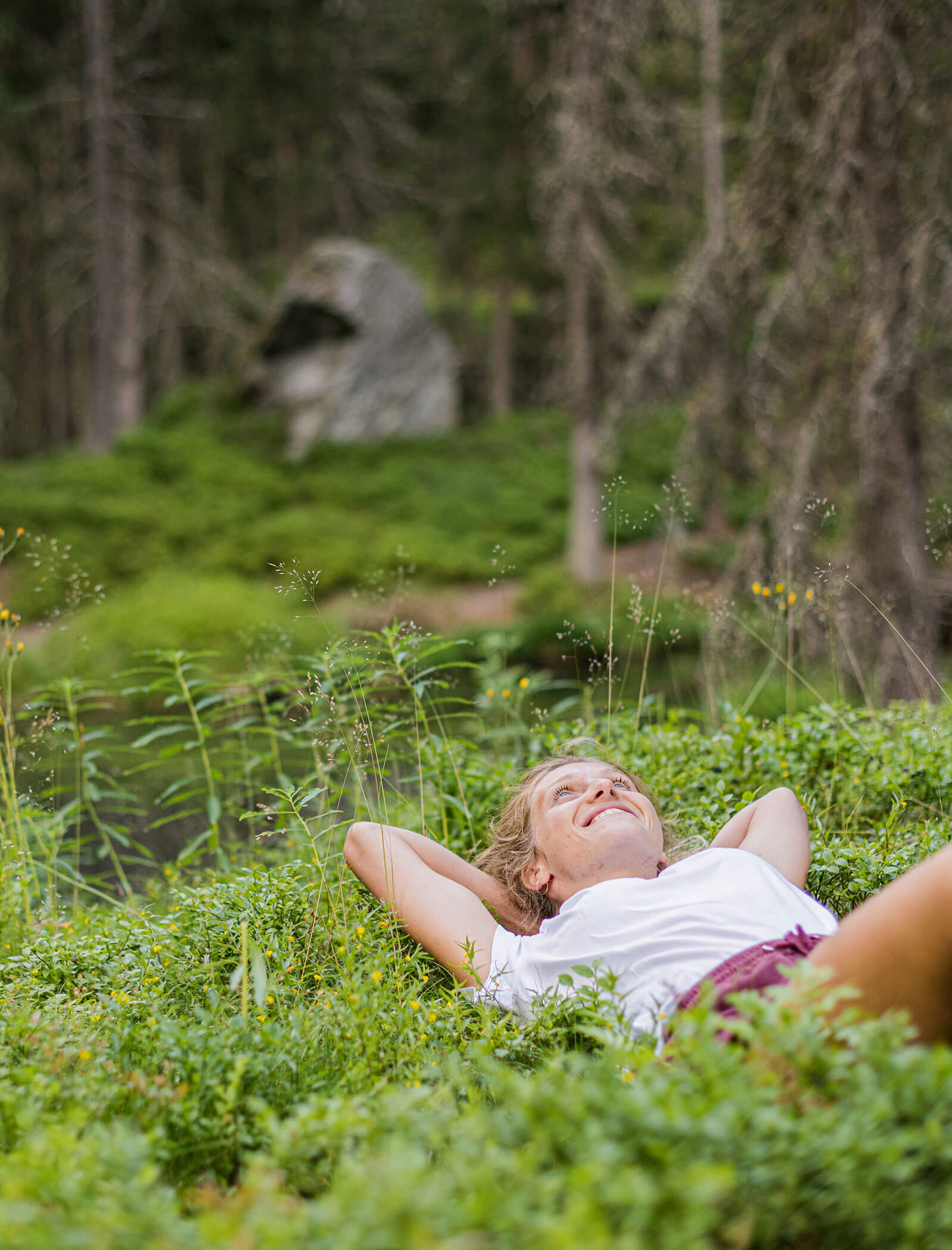 Auf dem Rücken liegende Frau im grünen Gras, lächelnd, mit einem Wald und einem großen Felsen im Hintergrund. - Hotel Moosmair
