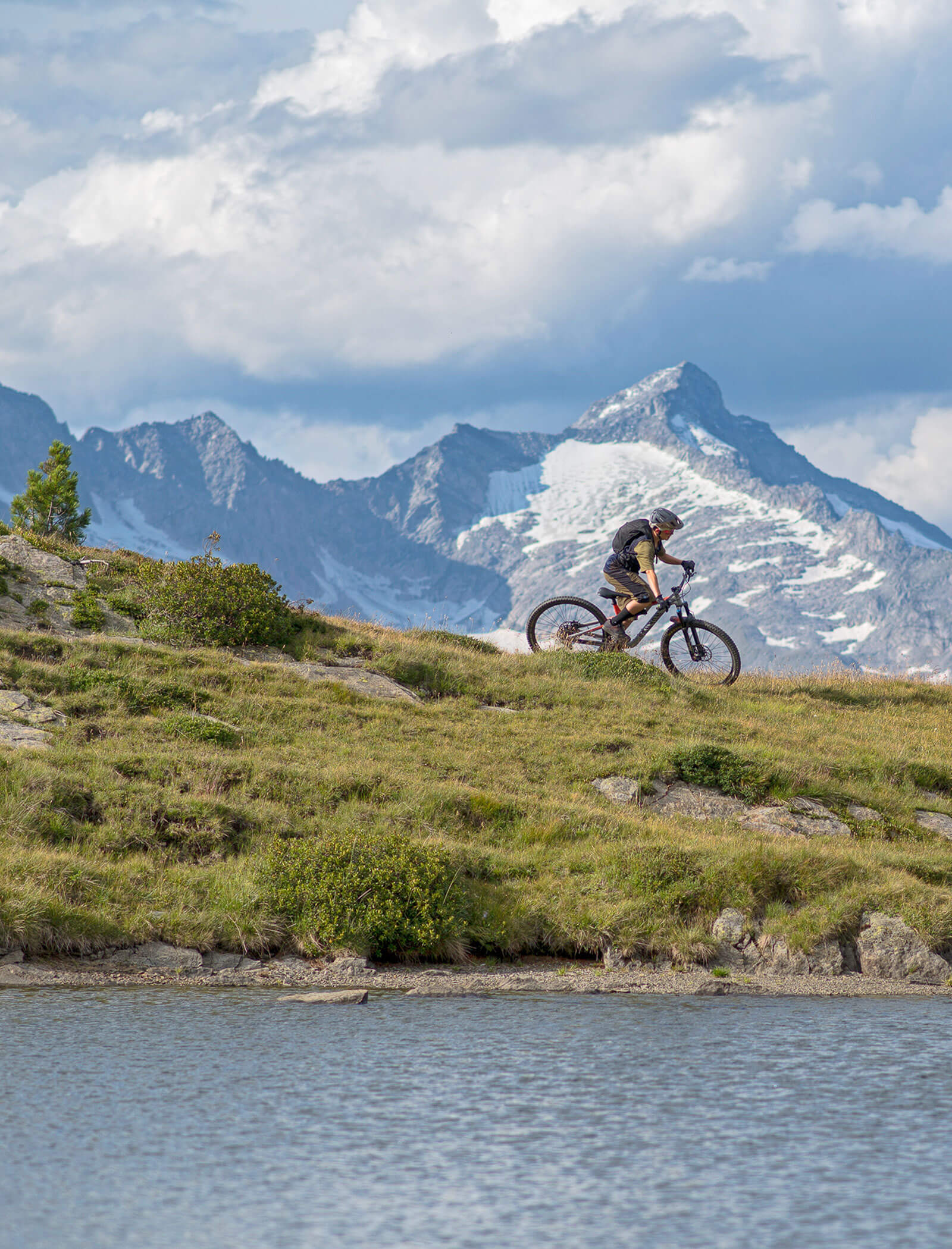 Ein Radfahrer fährt mit seinem Mountainbike auf einem grasbewachsenen Hügel mit verschneiten Gipfeln und einem See im Hintergrund. - Hotel Moosmair