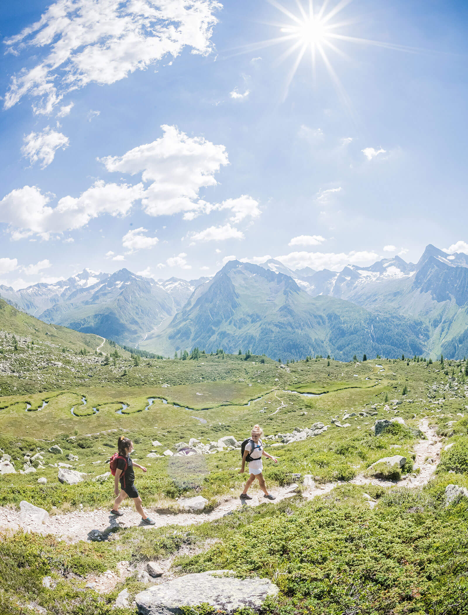 Zwei Wanderer gehen bei strahlendem Sonnenschein einen Bergpfad entlang, mit malerischen Alpenbergen im Hintergrund. - Hotel Moosmair