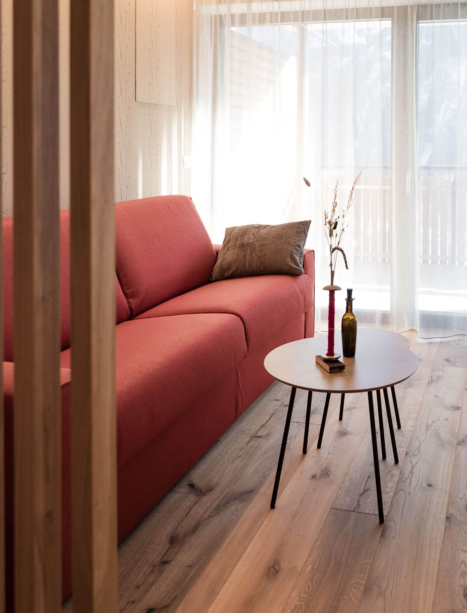 Ein rotes Sofa und ein runder Couchtisch aus Holz in einem sonnendurchfluteten Wohnzimmer mit Holzboden und transparenten Vorhängen. - Hotel Moosmair