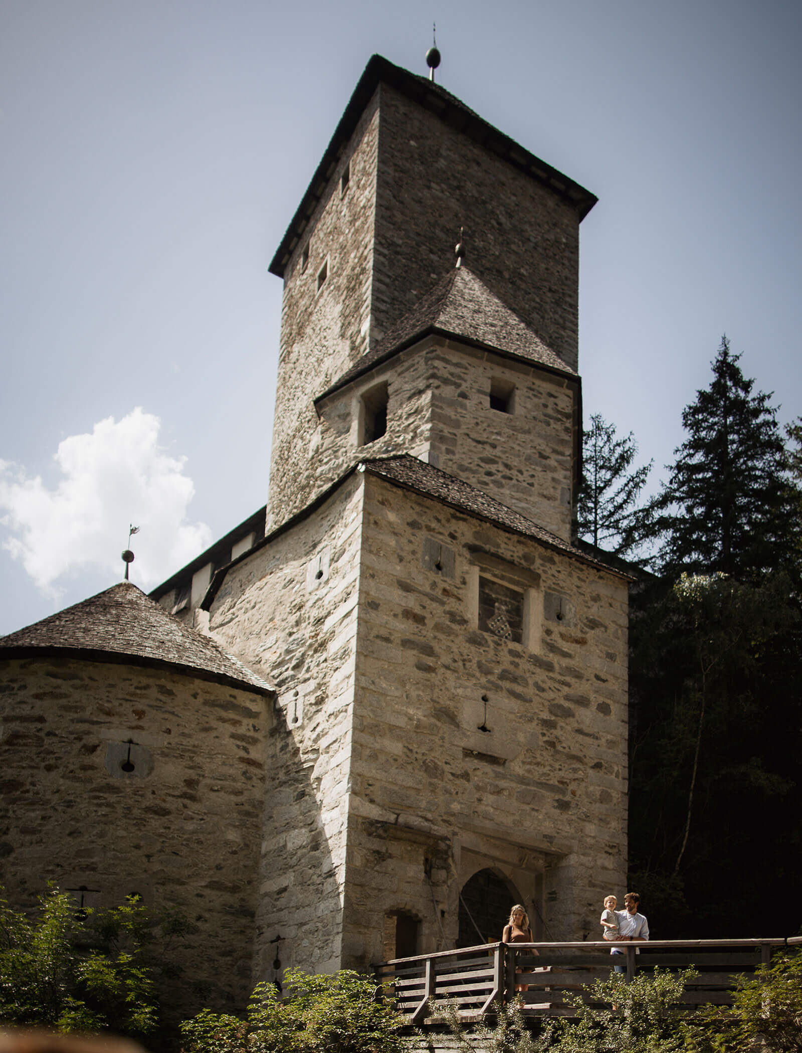 Zwei Personen stehen auf einer Holzbrücke vor einem hohen, alten Steinturm, der von Bäumen umgeben ist. - Hotel Moosmair