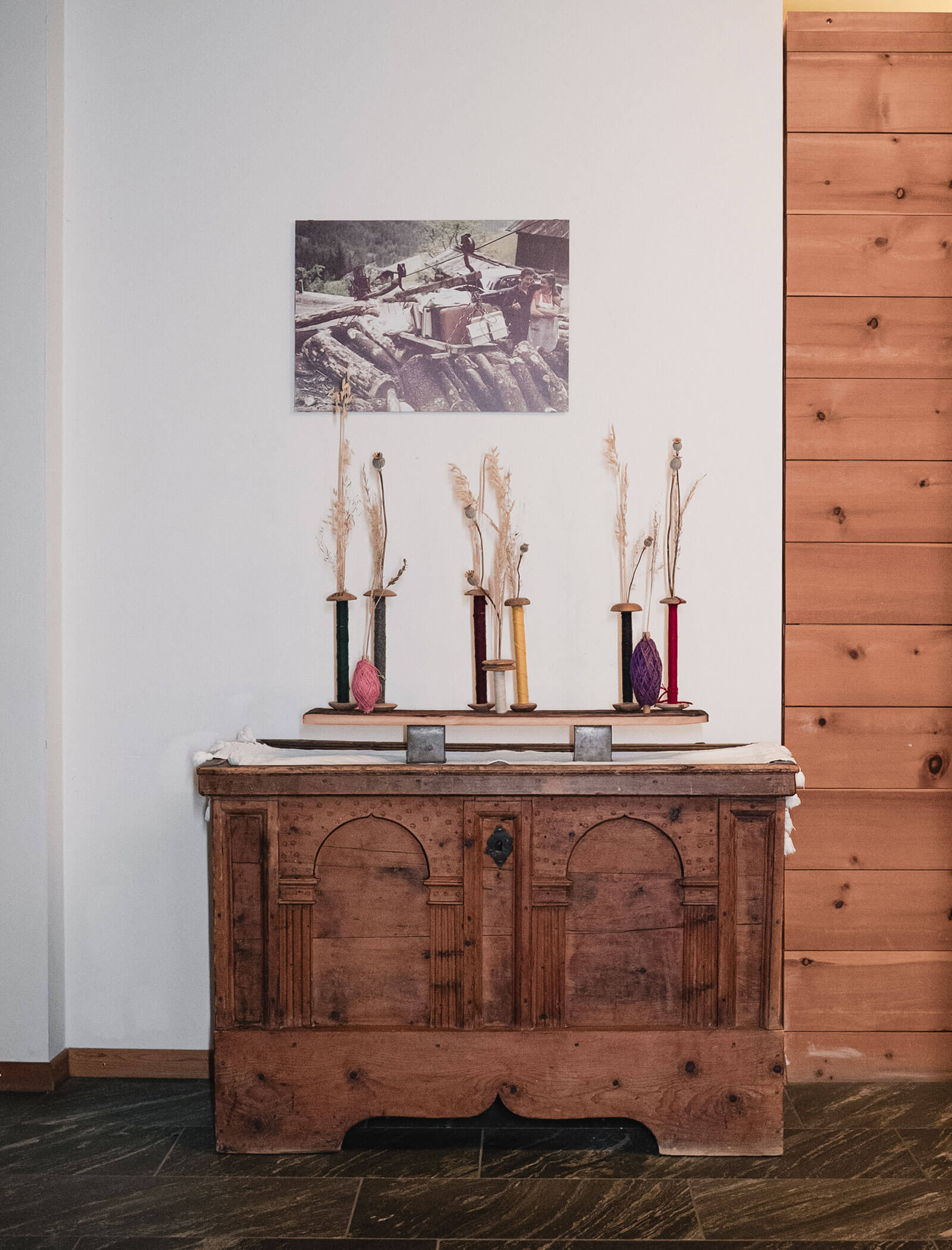Eine Holztruhe mit getrockneten Pflanzen, Garnspulen und einem alten Foto an der Wand darüber. - Hotel Moosmair