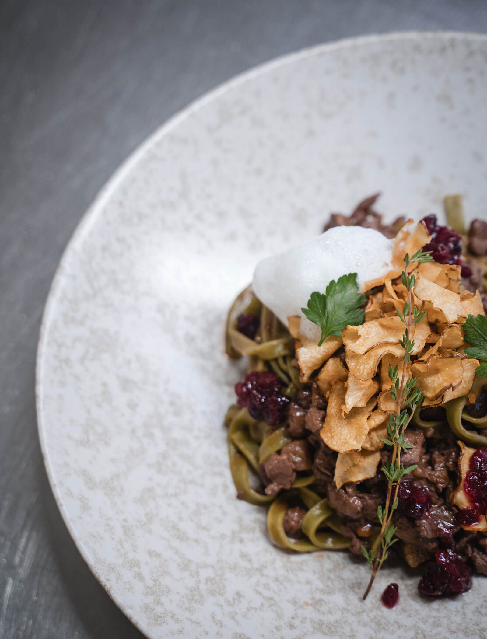 Tagliatelle-Nudeln mit Fleisch, Beilage, Schaum, Kräutern und knusprigen Chips auf einem gesprenkelten weißen Teller. - Hotel Moosmair