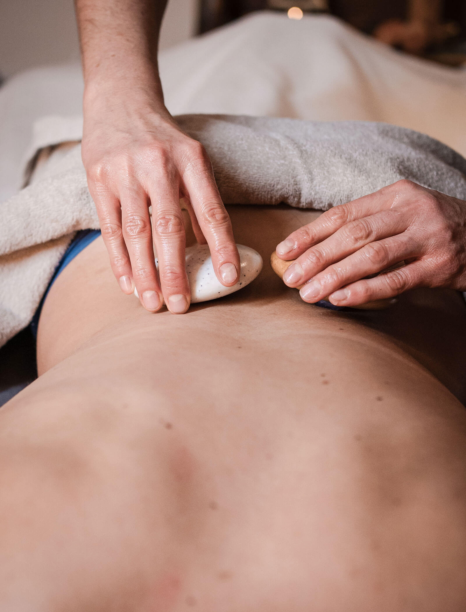 Hände, die eine Hot-Stone-Massage auf dem nackten Rücken einer Person durchführen, die teilweise von einem Handtuch bedeckt ist. - Hotel Moosmair