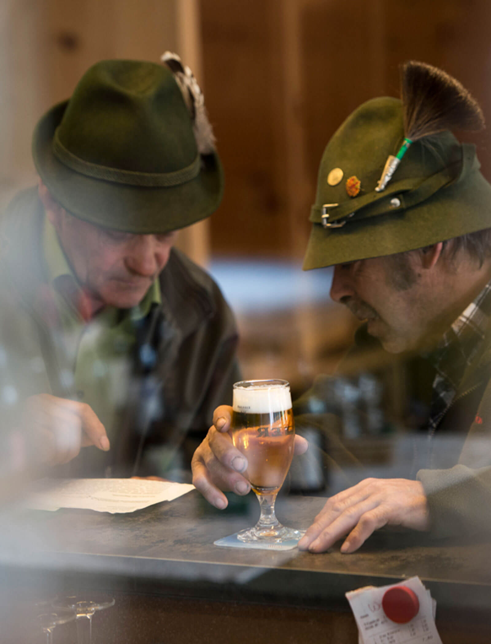 Zwei Männer mit grünen Hüten und Federn sitzen an einem Tisch, lesen eine Zeitung und haben ein Glas Bier vor sich stehen. - Hotel Moosmair