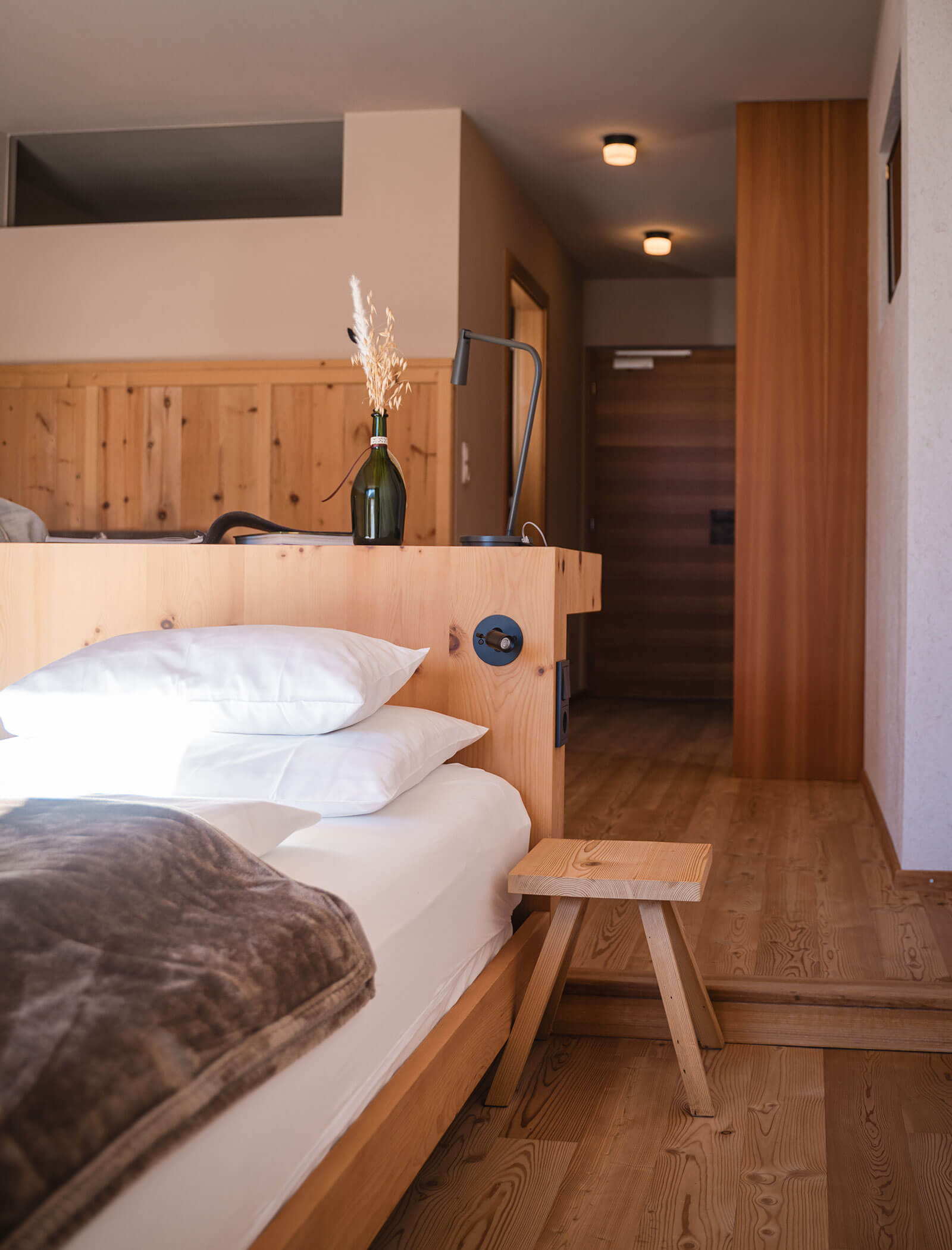 Minimalistisches Schlafzimmer aus Holz mit einem ordentlich gemachten Bett, Kissen, einem kleinen Hocker und einer Vase mit Trockenblumen. - Hotel Moosmair