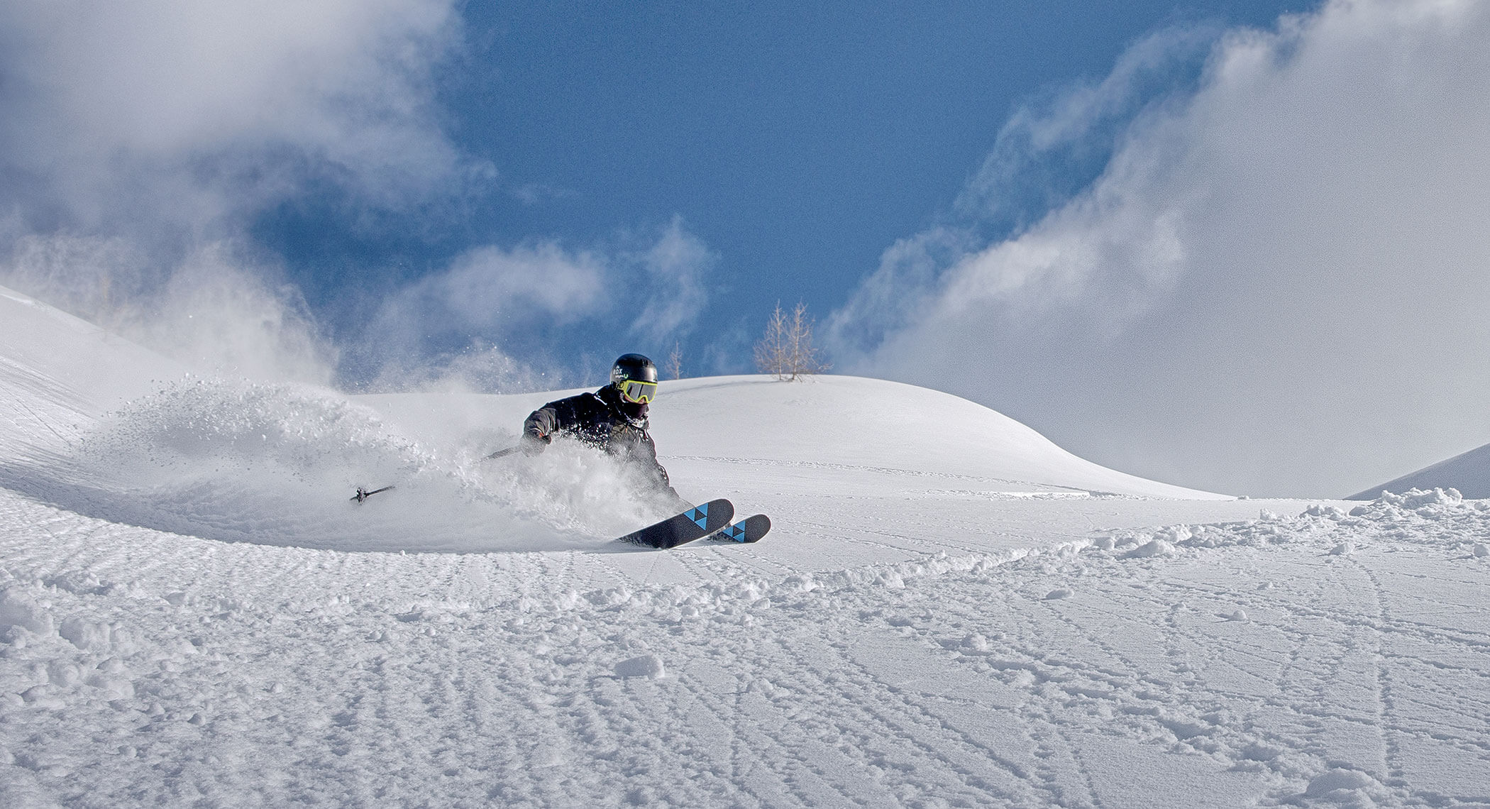 Skifahrer mit Skibrille macht eine scharfe Kurve auf frischem Pulverschnee unter einem blauen Himmel mit vereinzelten Wolken. - Hotel Moosmair