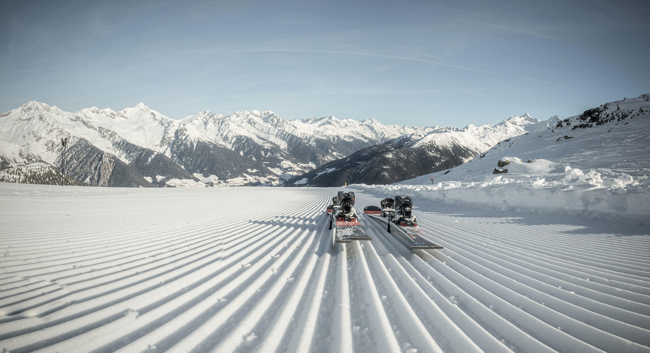 Zwei Paar Skier ruhen auf frisch präpariertem Schnee mit verschneiten Bergen im Hintergrund unter einem klaren Himmel. - Hotel Moosmair