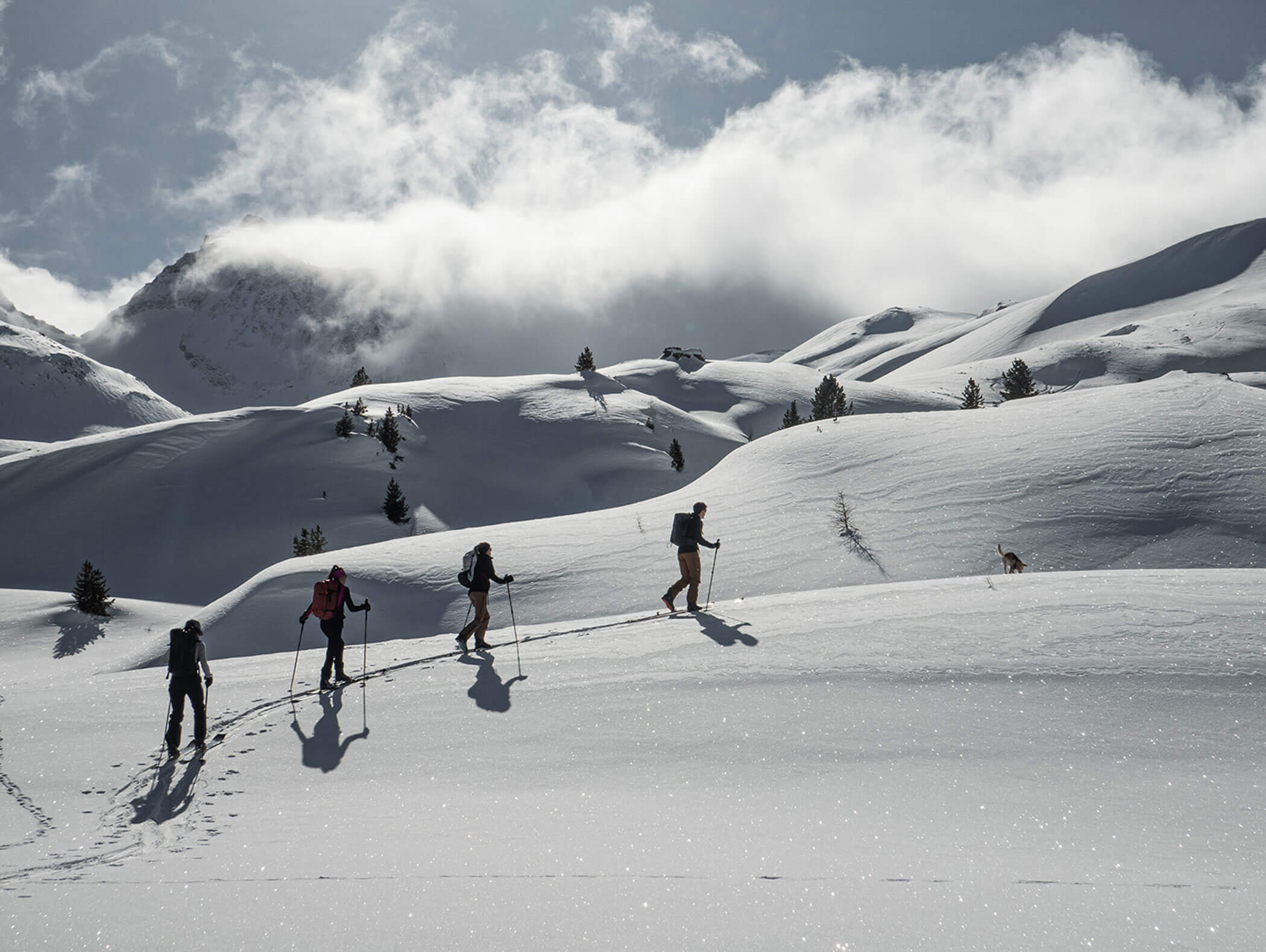 Vier Personen wandern bei bewölktem Himmel bergauf durch verschneite Berge und hinterlassen dabei Fußspuren im Schnee. - Hotel Moosmair