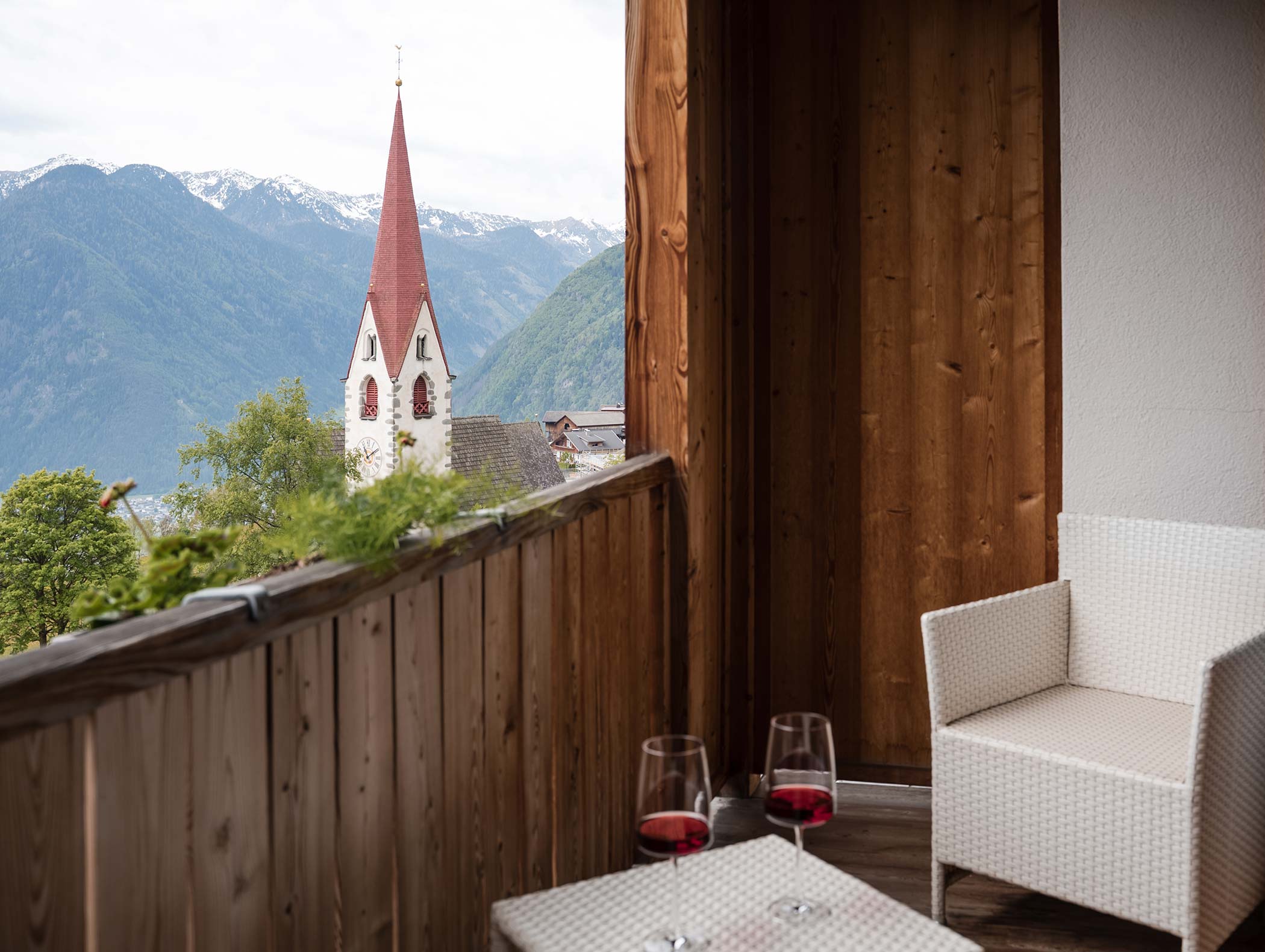 Von einem Balkon mit zwei Gläsern Rotwein blickt man auf einen Kirchturm und die Berglandschaft. - Hotel Moosmair