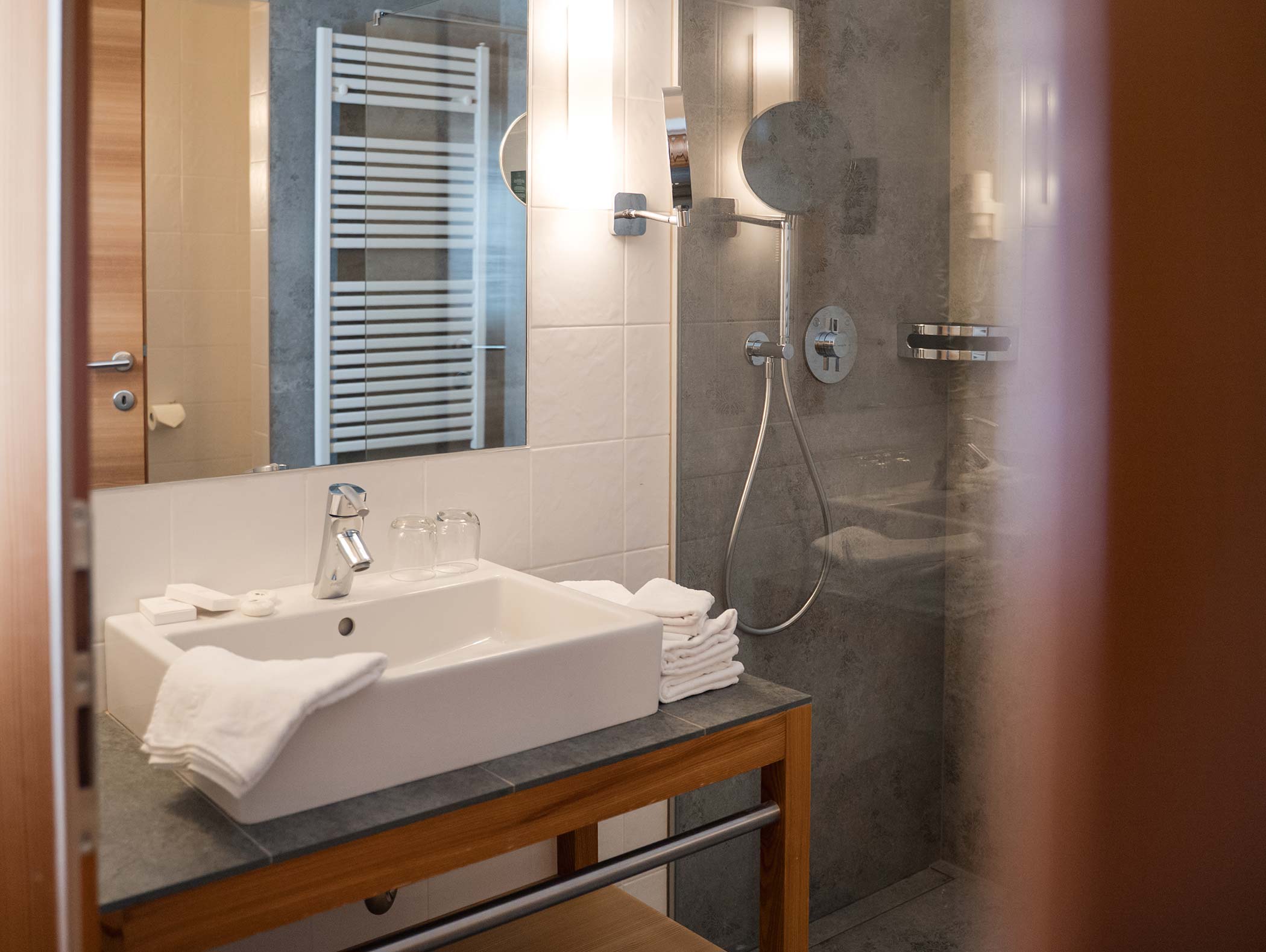 Modernes Badezimmer mit Waschbecken, Spiegel, Handtüchern, Gläsern und einer wandmontierten Dusche in einer Glasabtrennung. - Hotel Moosmair