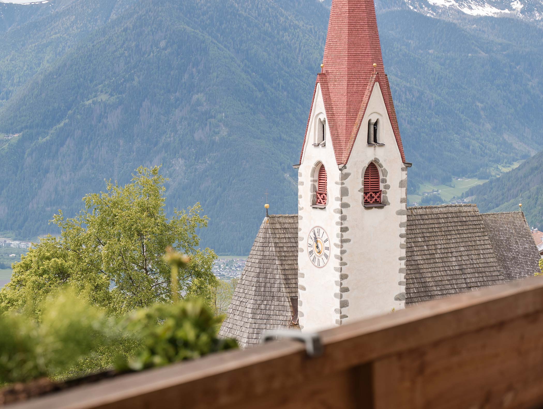 Ein Kirchturm mit einer Uhr steht an einem bewölkten Tag vor Bergen und Grünanlagen. - Hotel Moosmair