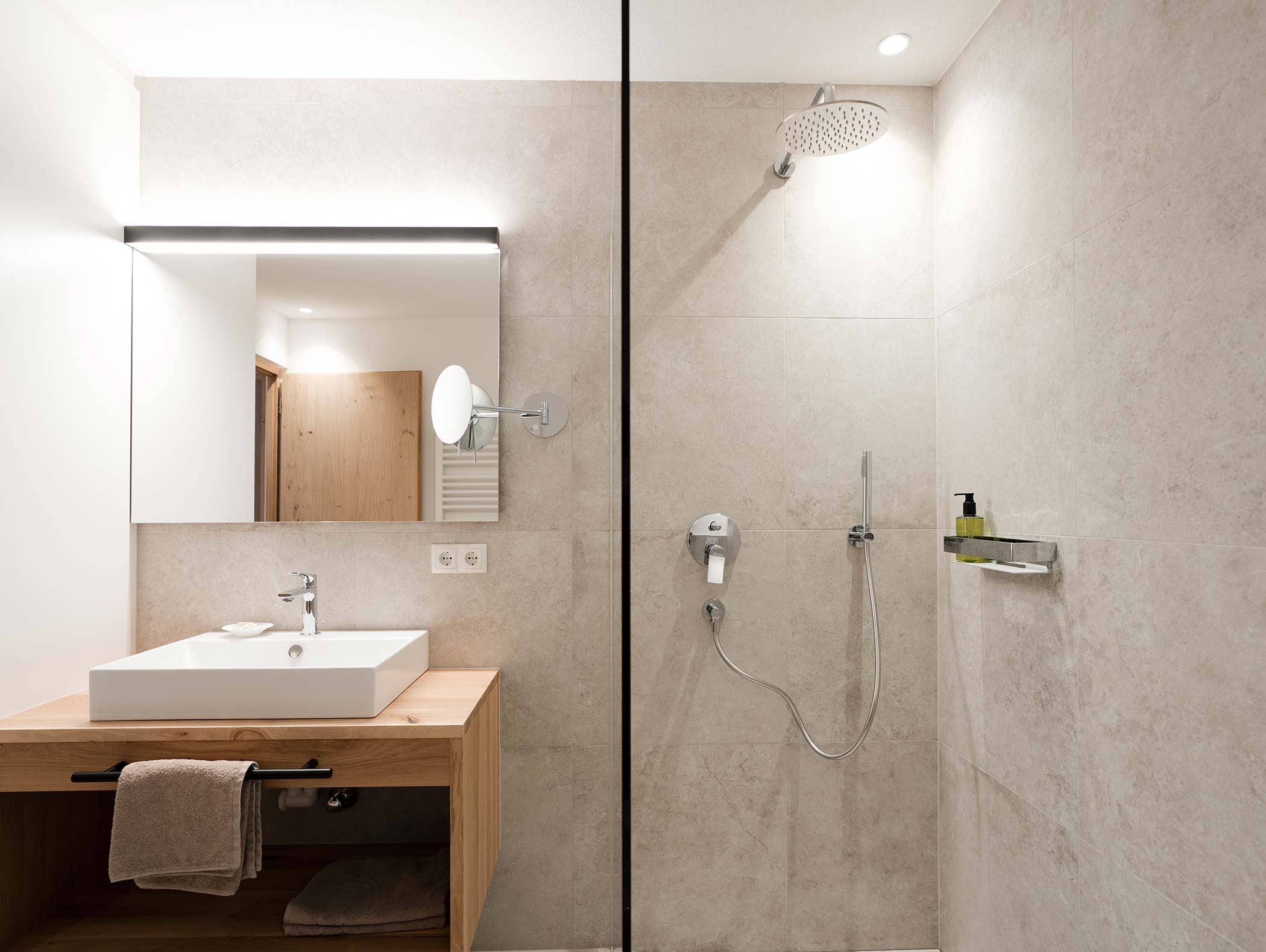 Modernes Badezimmer mit einem großen Spiegel, einem Waschbecken auf einem hölzernen Waschtisch und einer begehbaren Dusche mit Regenduschkopf. - Hotel Moosmair
