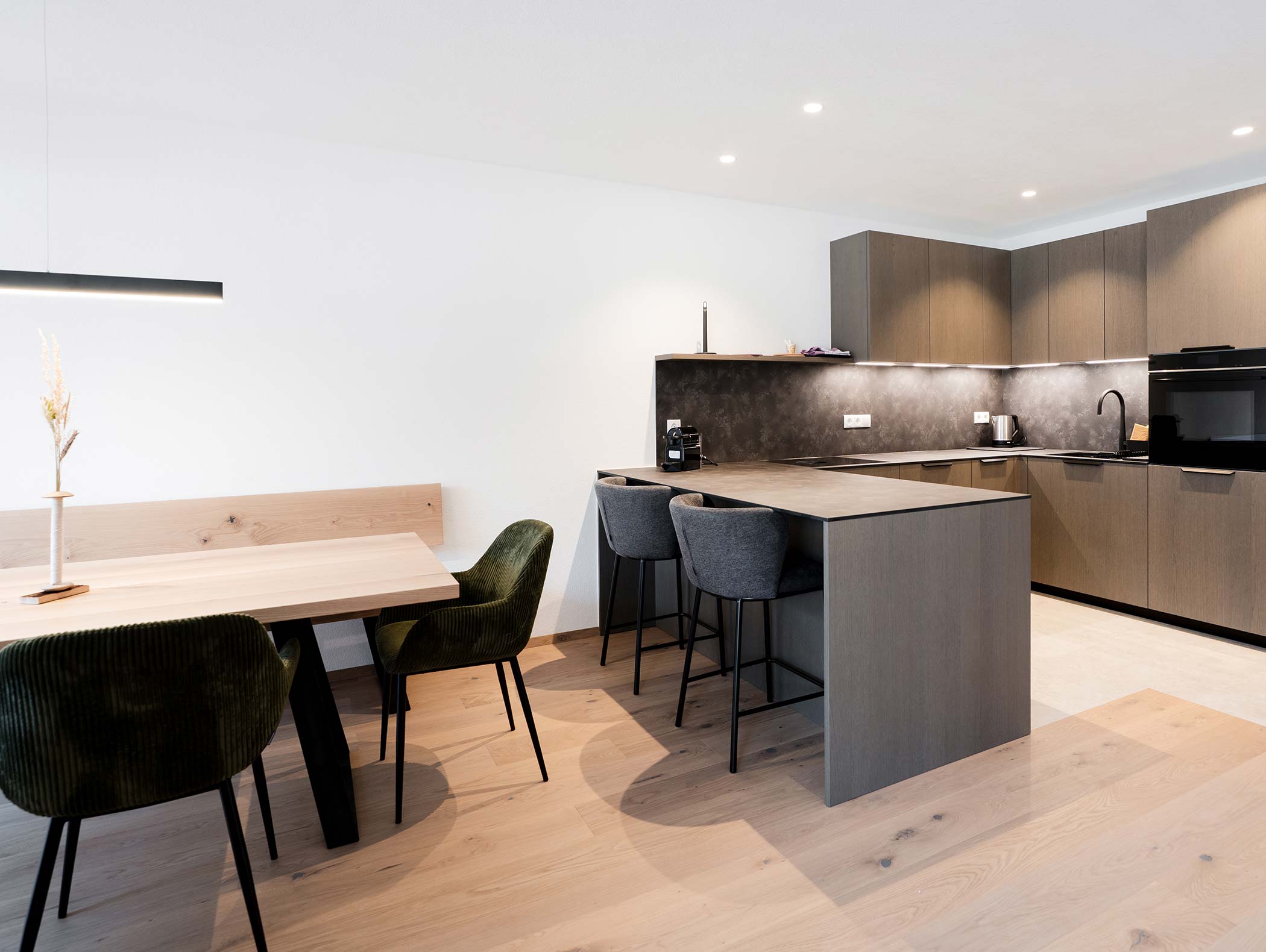 Moderne Küche mit Holzböden, Insel mit Barhockern und Esstisch mit grünen Stühlen unter Hängelampen. - Hotel Moosmair