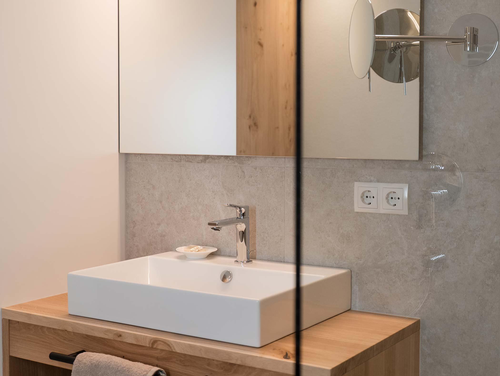 Modernes Waschbecken mit quadratischem Becken, Wandspiegel und Arbeitsplatte aus Holz mit Handtuchhalter. - Hotel Moosmair
