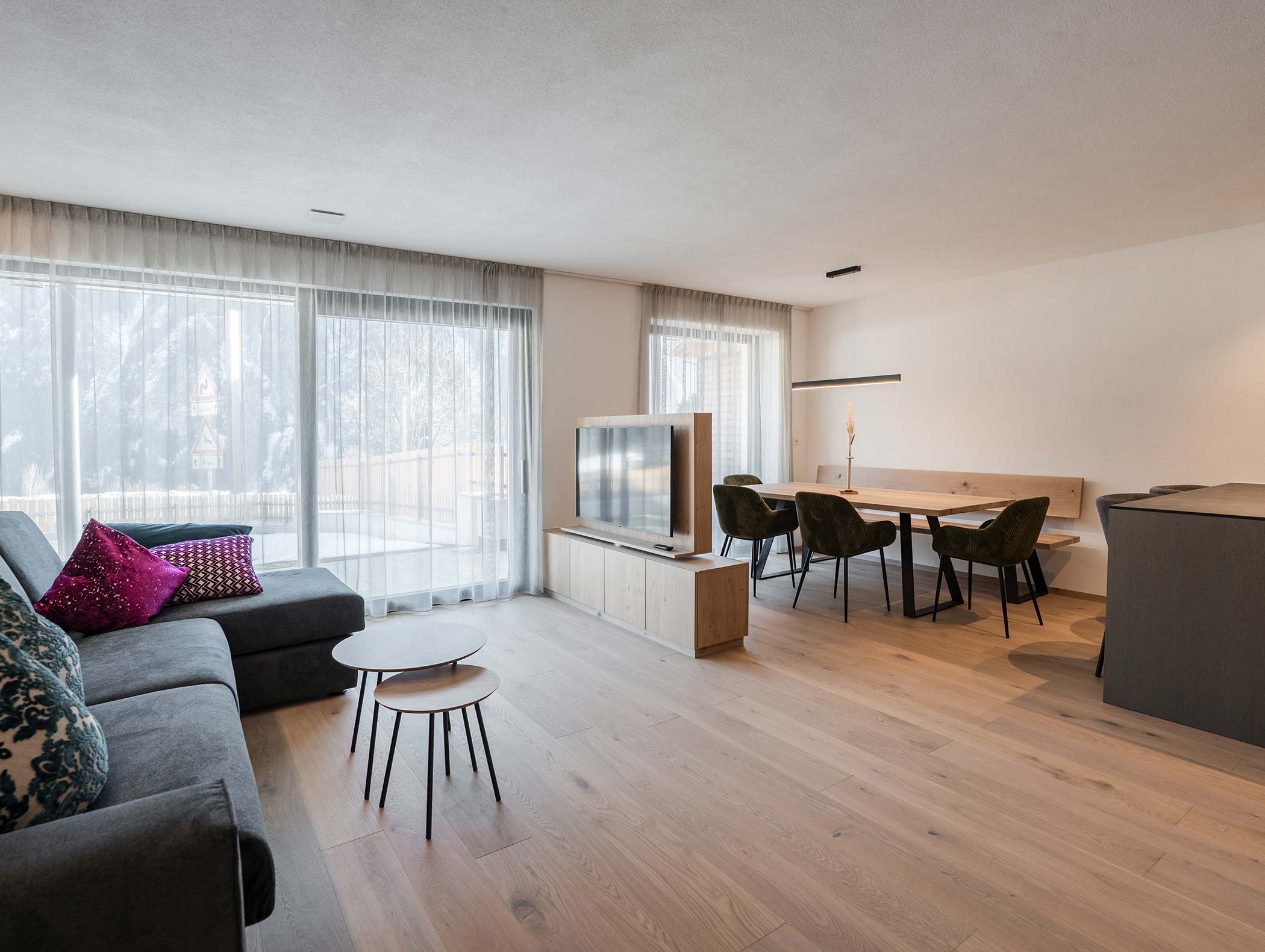 Modernes Wohn- und Esszimmer mit großen Fenstern, einem grauen Sofa, TV und Holzfußboden. - Hotel Moosmair