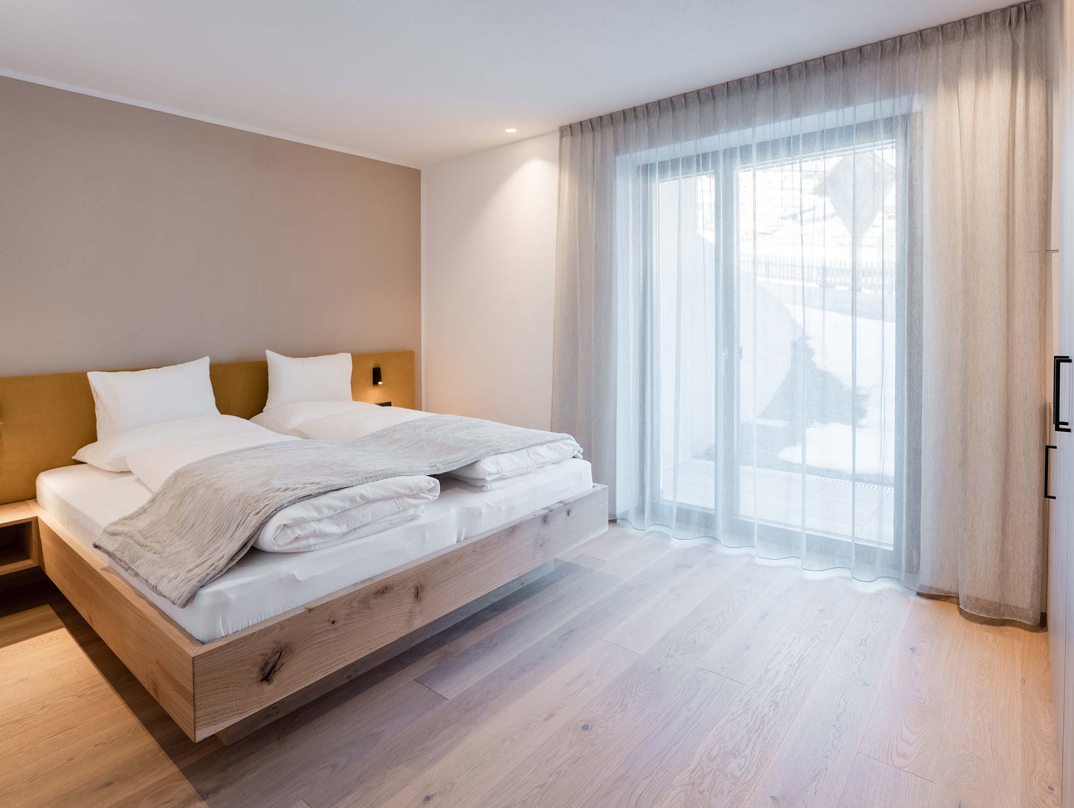 Minimalistisches Schlafzimmer mit einem Doppelbett, hellem Holzboden und transparenten Vorhängen vor einem großen Fenster. - Hotel Moosmair