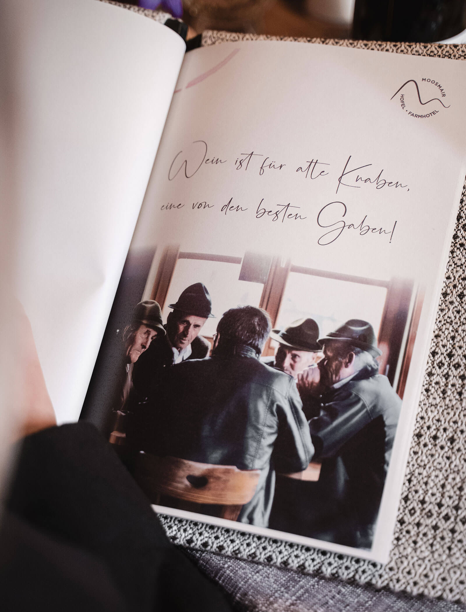 Eine Gruppe älterer Männer mit Hüten sitzt an einem Holztisch in der Nähe eines Fensters und unterhält sich, von hinten fotografiert. - Hotel Moosmair