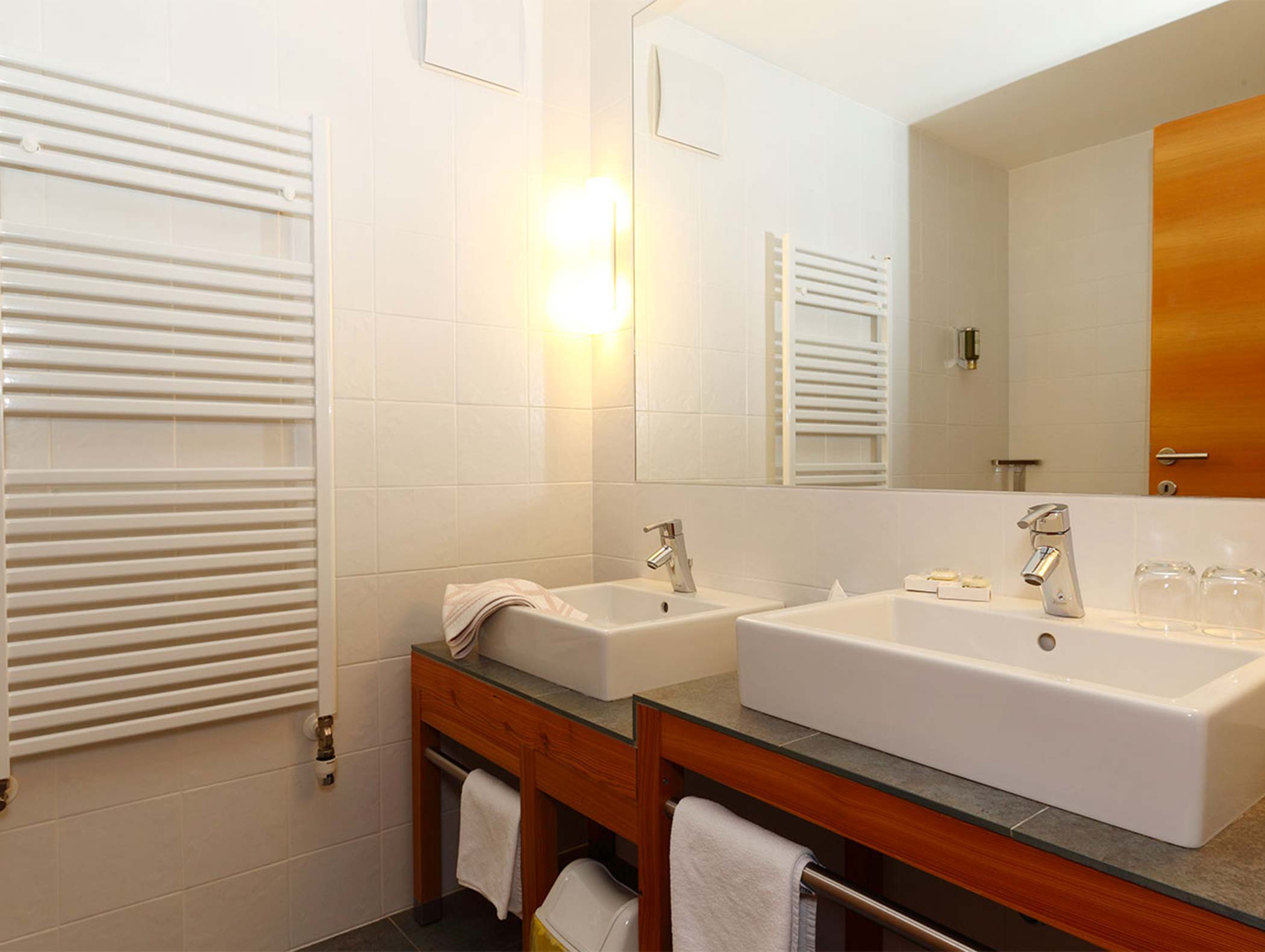 Modernes Badezimmer mit zwei weißen Waschbecken, einem hölzernen Waschtisch, Handtüchern und einem großen Spiegel über dem Tresen. - Hotel Moosmair