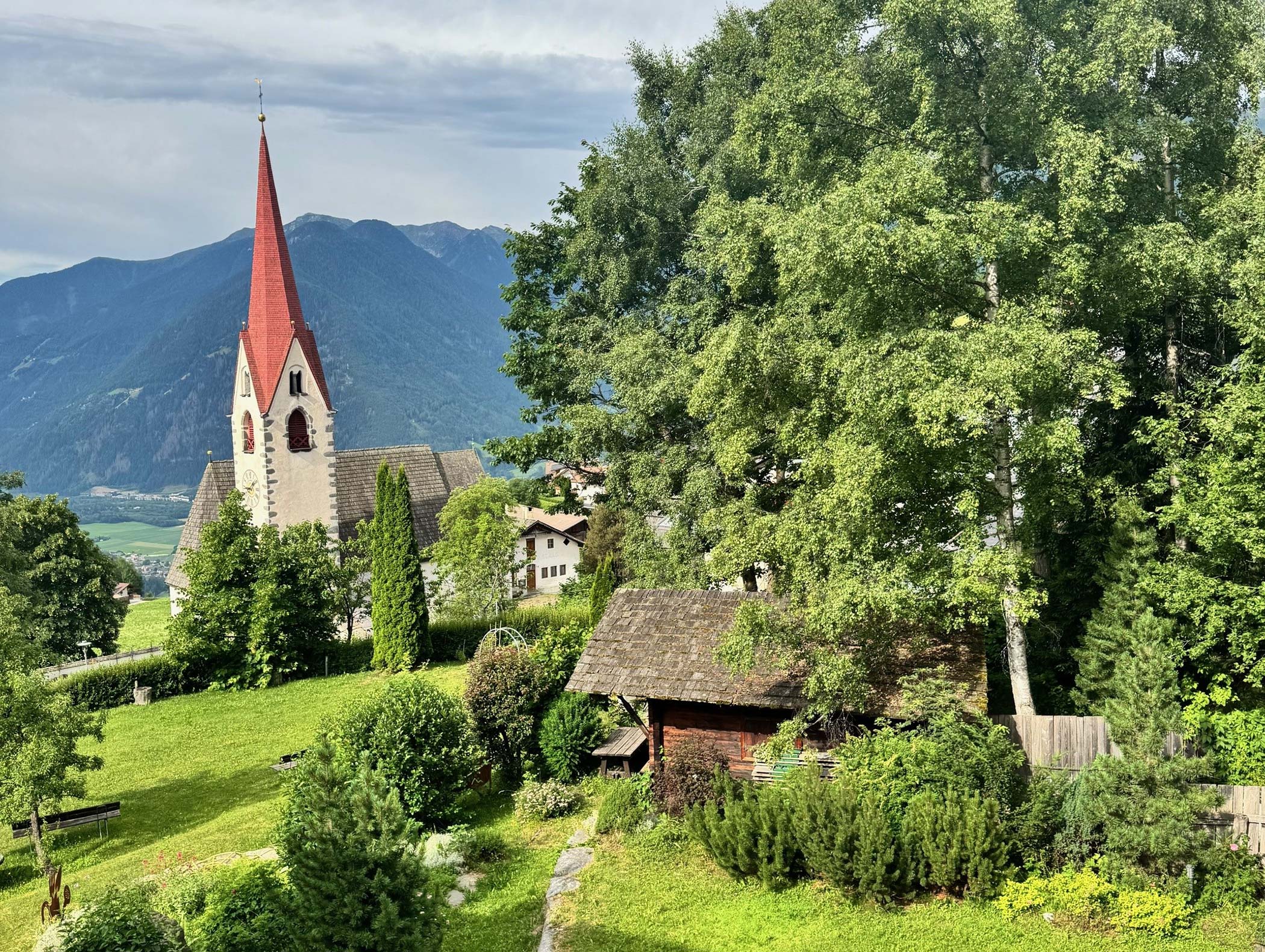 Eine rot gedeckte Kirche und Holzhäuser liegen zwischen grünen Bäumen und Bergen im Hintergrund. - Hotel Moosmair