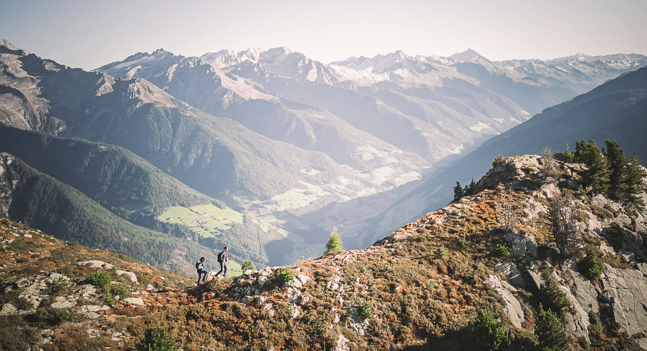 Zwei Wanderer wandern entlang eines Bergkamms mit malerischen, sonnenbeschienenen Tälern und schneebedeckten Gipfeln im Hintergrund. - Hotel Moosmair