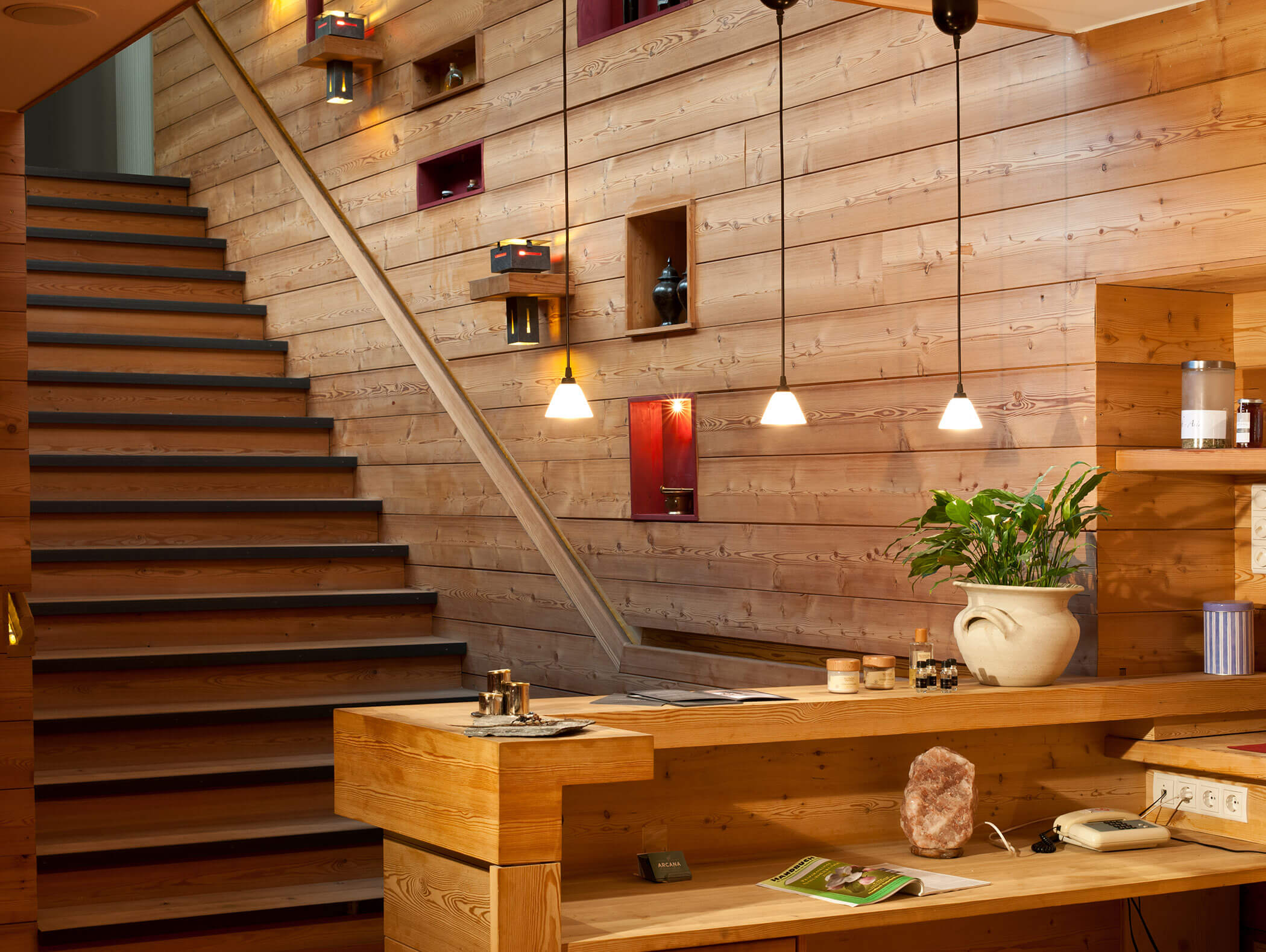 Moderner hölzerner Empfangsbereich mit Treppe, Hängelampen und Dekorationsartikeln in Regalen und auf der Theke. - Hotel Moosmair