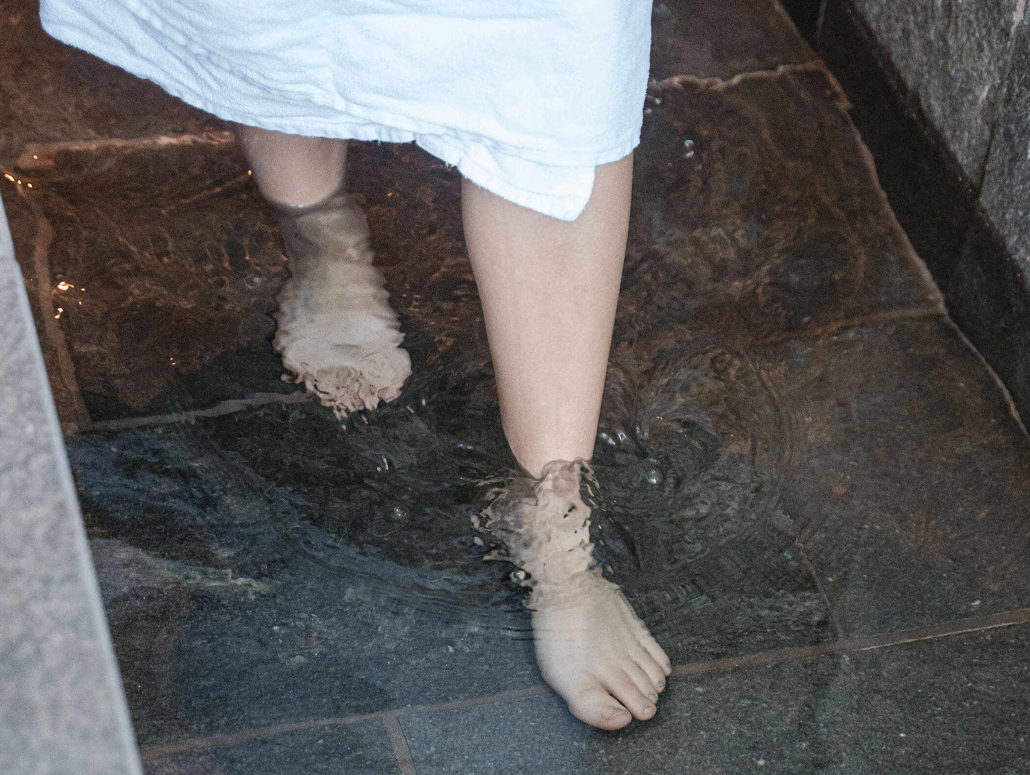 Person in einem weißen Handtuch, die barfuß in flachem Wasser auf einem Steinboden steht. - Hotel Moosmair