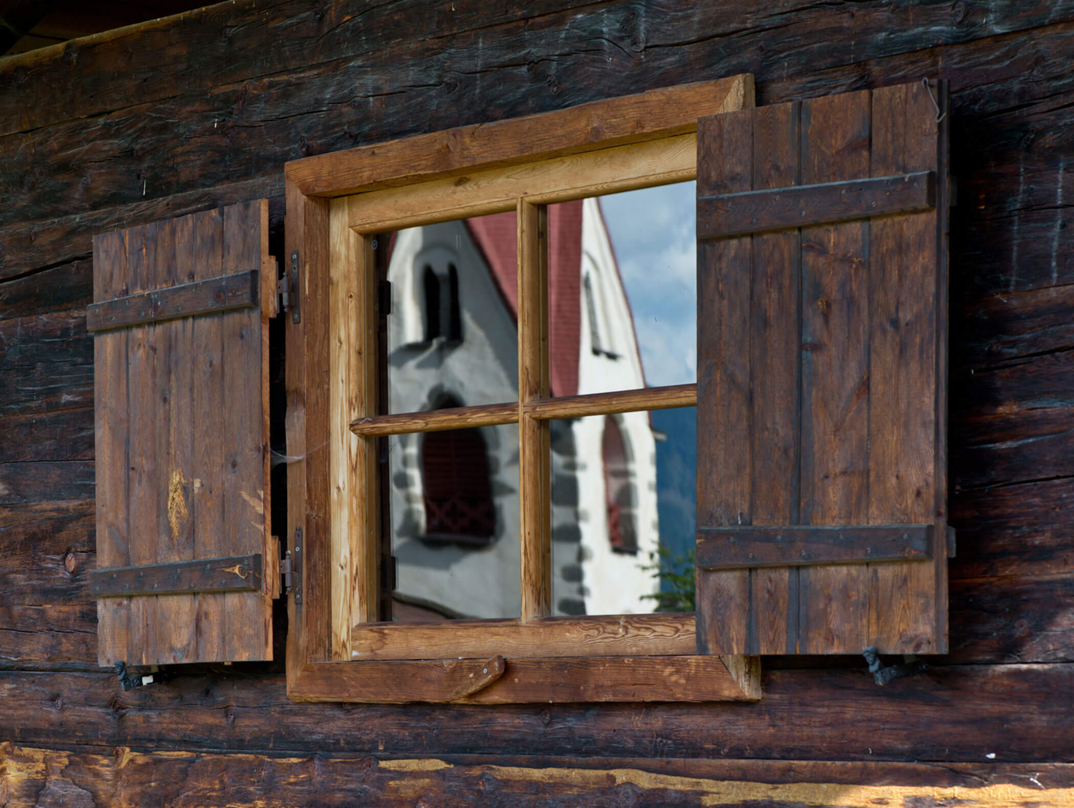 Ein Holzfenster mit offenen Fensterläden spiegelt eine weiße Kirche mit rotem Dach und Bogenfenstern wider. - Hotel Moosmair