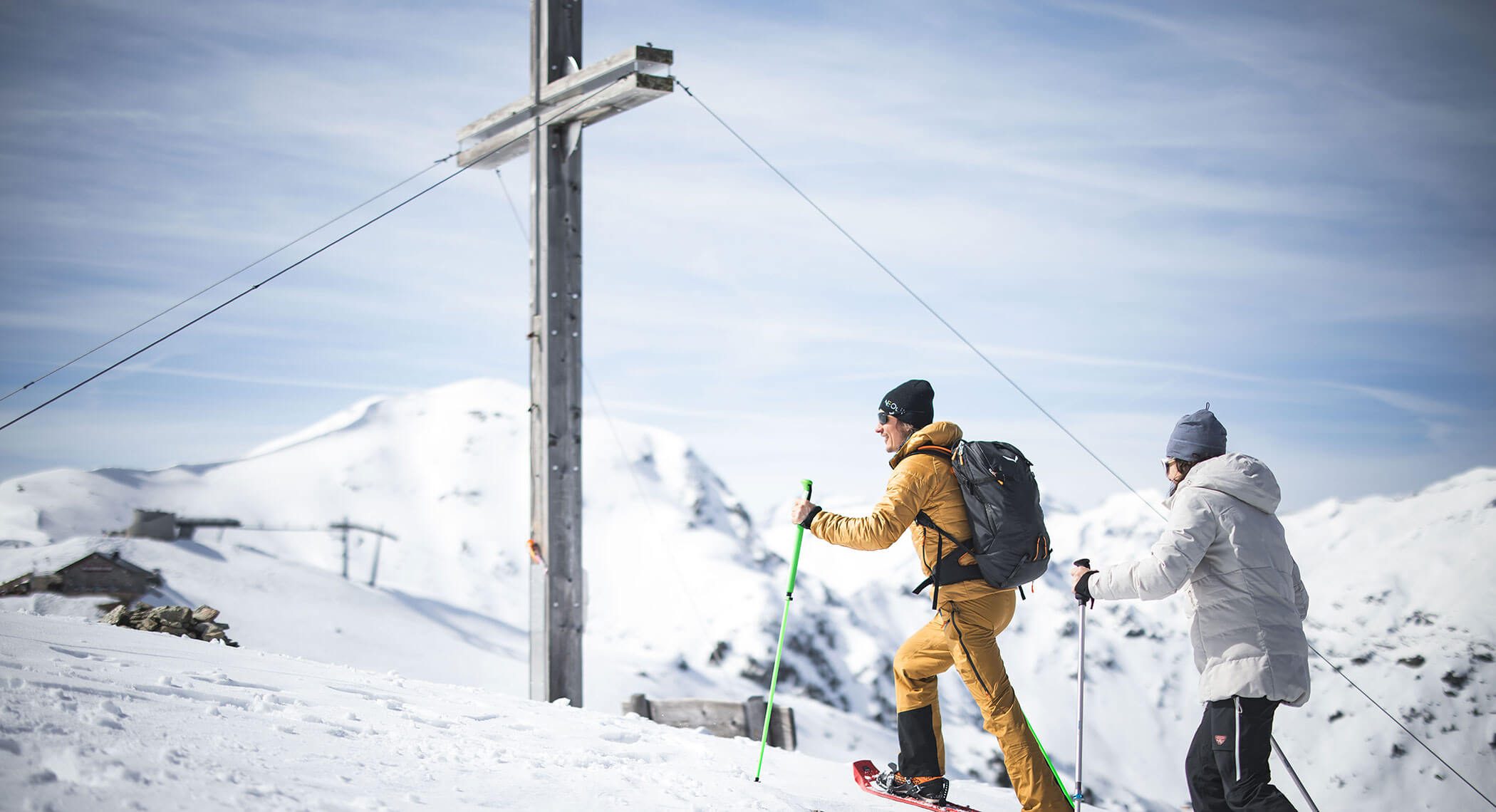 Zwei Personen mit Rucksäcken wandern auf einen verschneiten Berg in der Nähe eines großen Kreuzes unter einem bewölkten Himmel. - Hotel Moosmair