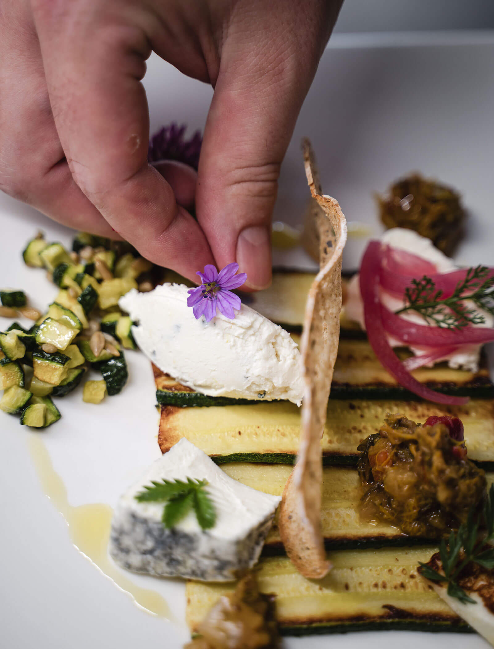 Eine Hand platziert eine lila Blume auf einem Gourmetteller mit Käse, gegrillten Zucchini und Beilagen auf einem weißen Teller. - Hotel Moosmair