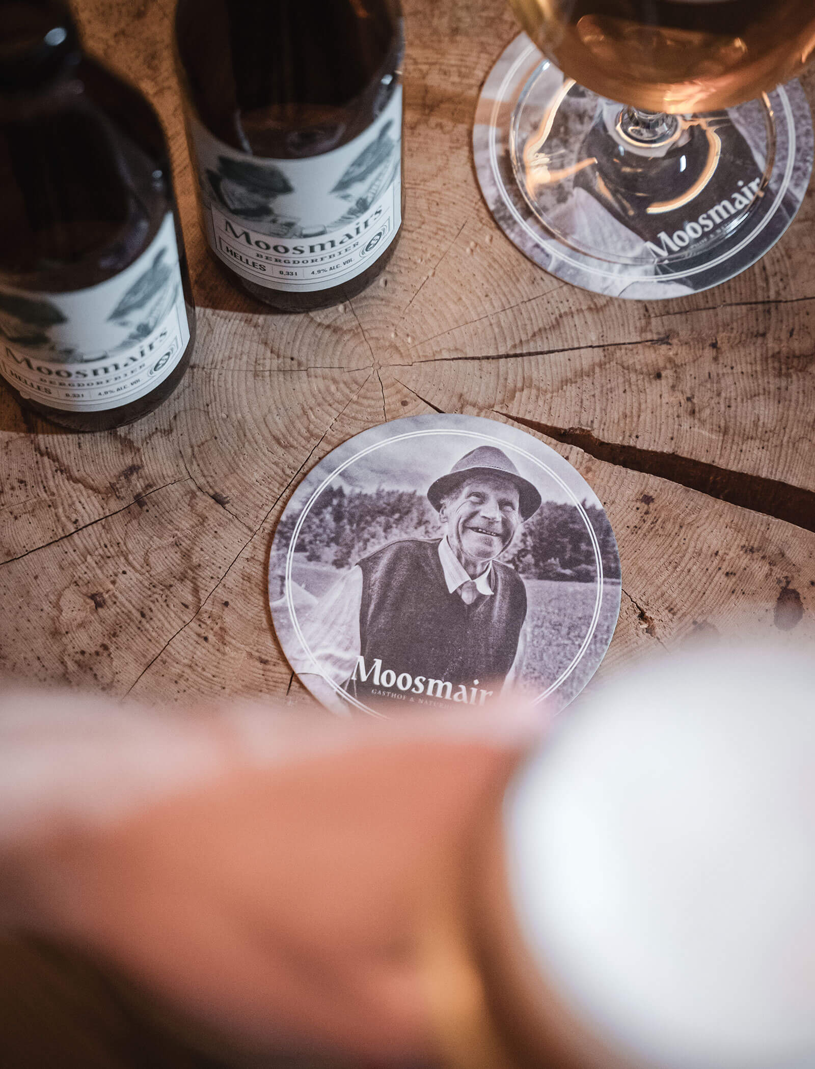 Ein runder Untersetzer mit einem Foto eines älteren Mannes und Moosmair, umgeben von Getränken auf einem Holztisch. - Hotel Moosmair
