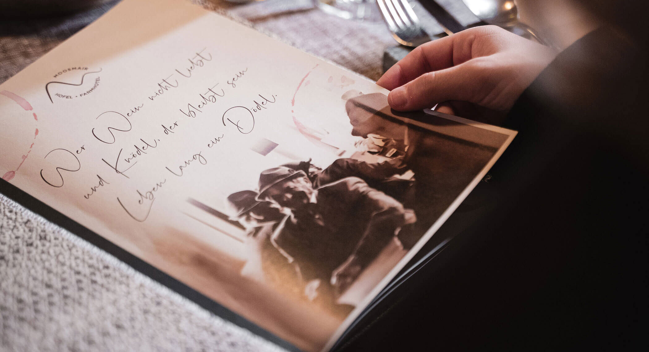 Eine Person hält eine Speisekarte mit deutschem Text und ein altes Foto von Menschen, die zusammensitzen. - Hotel Moosmair
